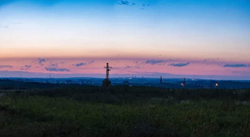 Panorama in der blauen Stunde: ein rosa Wolkenband über der dunklen Ebene, in der Mitte ein Strommast als Silhouette, am Horizont erste Lichter
