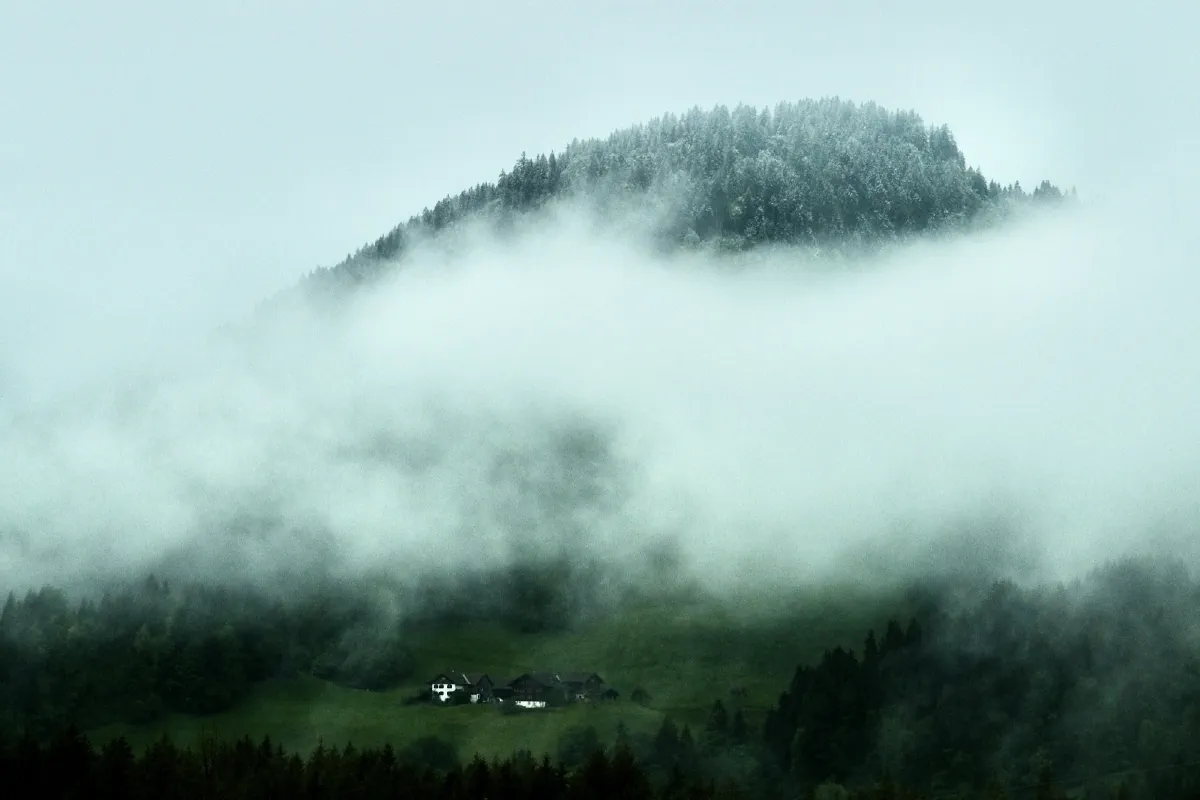 Bewaldeter Bergrücken, ein Wolkenband zieht quer darüber, darunter liegt ein einzelner Hof am Hang