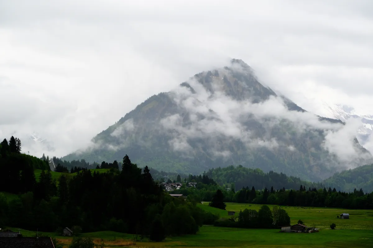 Markanter Berggipfel, halb in Wolken, darunter ein Dorf zwischen Wiesen und Nadelwald