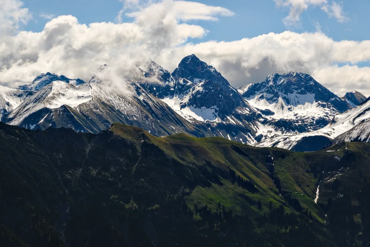 Schneebedeckte Gipfelkette, davor ein dunkler, grün bewachsener Bergrücken im Schatten