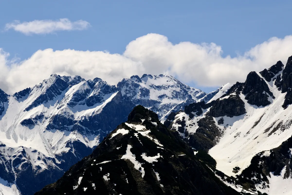 Verschneite Gipfel hinter einem dunklen Felsgrat, darüber Quellwolken