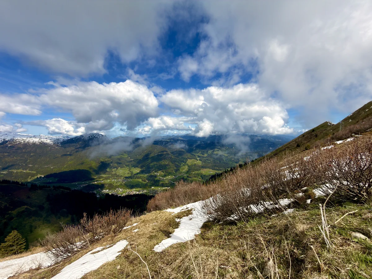 Blick von einem Hang mit Schneeresten hinunter ins Tal, Wolken ziehen über die gegenüberliegende Bergkette