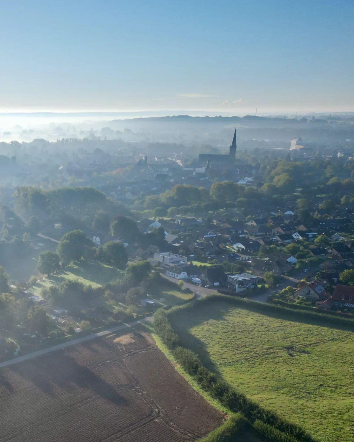 Luftaufnahme einer Ortschaft im Morgennebel, der Kirchturm ragt aus dem Dunst, davor Wiesen und ein gestreifter Acker