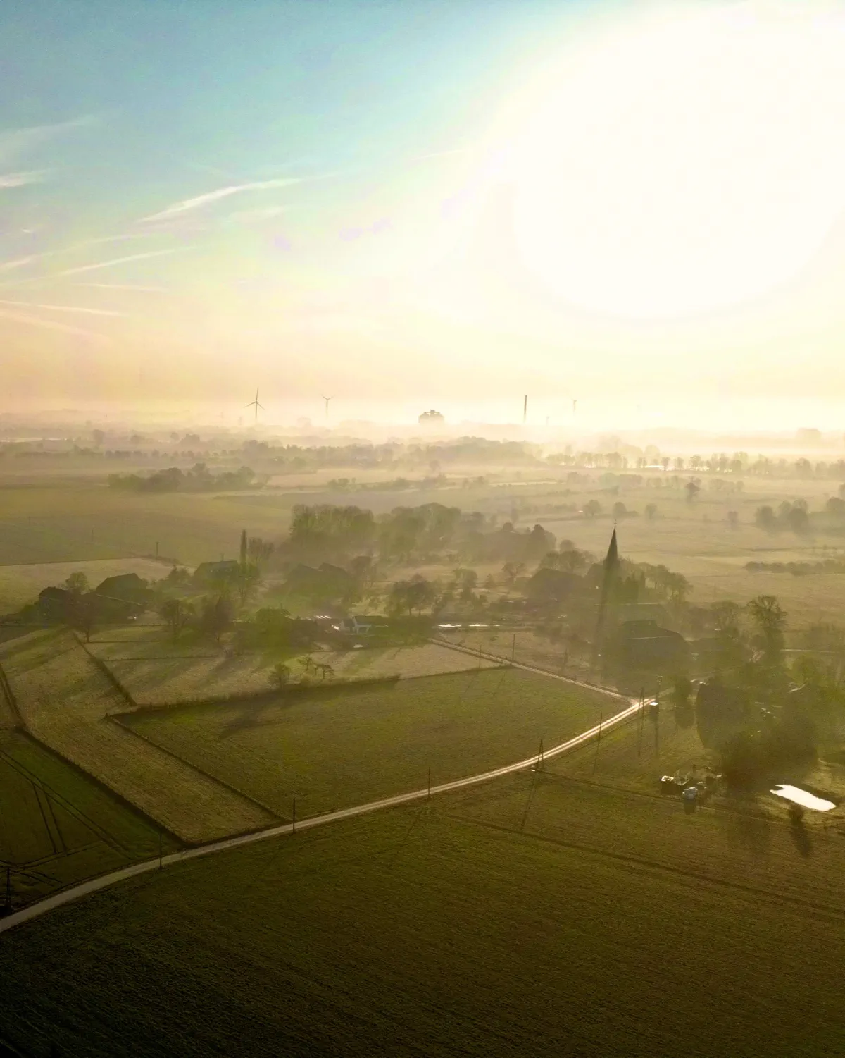 Luftaufnahme eines Dorfes im Gegenlicht, Nebel liegt über den umliegenden Feldern, am Horizont Windräder