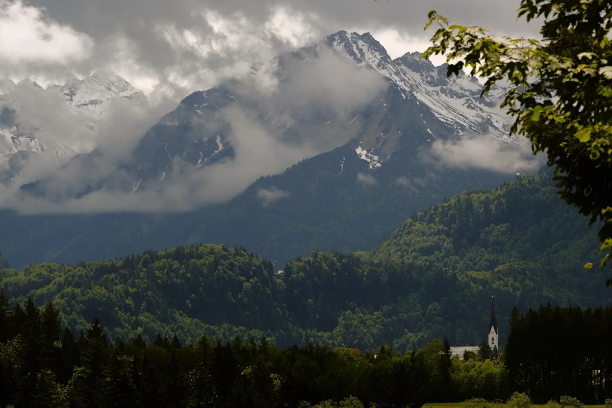 Bergkette mit Wolkenbändern über bewaldeten Hängen, im Tal ein Kirchturm
